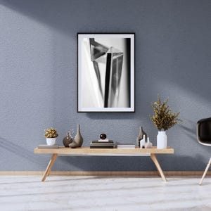 May include: A monochrome framed print with a geometric design hangs on a textured blue wall. A wooden coffee table displays decorative vases, a small potted plant, and books. A white vase holds dried flowers.