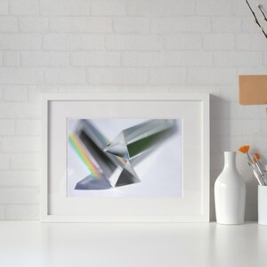 May include: A framed photograph featuring two clear glass prisms, one casting a rainbow spectrum. The image is set against a white background, with a white frame. A white vase and a cylindrical container with paintbrushes are in the foreground.