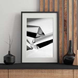 May include: Monochrome framed print of a geometric prism. The artwork is displayed on a black surface, with a black vase and a decorative vase to the right. The background includes wooden panels.