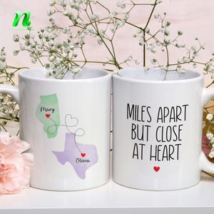 May include: Two white ceramic mugs. One mug features a green and purple map design with the names "Mary" and "Olivia" and a heart. The other mug has the text "MILES APART BUT CLOSE AT HEART" with a red heart.
