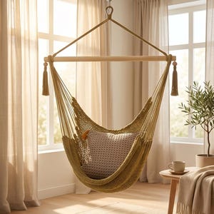 May include: A hanging chair made of olive green woven cord featuring a horizontal wooden spreader bar at the top, decorative tassels at each end, and a single square textured pillow in a neutral beige tone placed inside the seat.