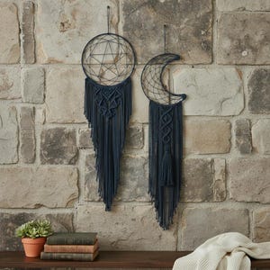 May include: Two dark blue macrame wall hangings, one a circle with a geometric design, the other a crescent moon shape. Both have long fringe and are suspended against a stone wall. Decorative home decor.