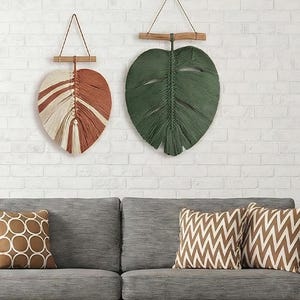 May include: Two leaf-shaped wall hangings. One is a mix of cream, brown, and rust-colored fibers, the other is green. Both are suspended from wooden bars with a leather cord. The leaves are mounted on a white brick wall.