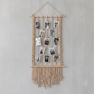 May include: A beige macrame photo display with a wooden dowel at the top and bottom. Vertical beige cords hang from the top dowel, holding small black and white photos. The sides feature braided macrame detailing, and the bottom has a fringe of knotted cords.