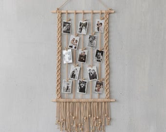 May include: A beige macrame photo display with a wooden dowel at the top and bottom. Vertical beige cords hang from the top dowel, holding small black and white photos. The sides feature braided macrame detailing, and the bottom has a fringe of knotted cords.