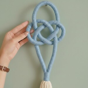 May include: A light blue macrame wall hanging featuring a Celtic knot design. The handmade piece is crafted from soft, woven material and includes a cream-colored tassel at the bottom. The wall decor is designed to be hung on a wall.