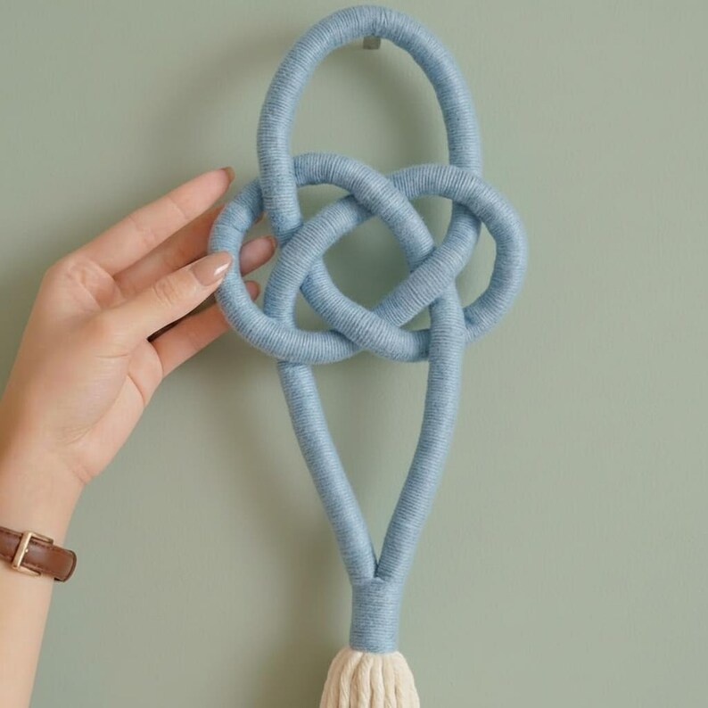 May include: A light blue macrame wall hanging featuring a Celtic knot design. The handmade piece is crafted from soft, woven material and includes a cream-colored tassel at the bottom. The wall decor is designed to be hung on a wall.