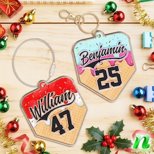 May include: Two personalized keychains shaped like baseball home plates. One keychain has the name "William" and the number "47" on a red and white design. The other keychain has the name "Benjamin" and the number "25" on a blue and pink design.