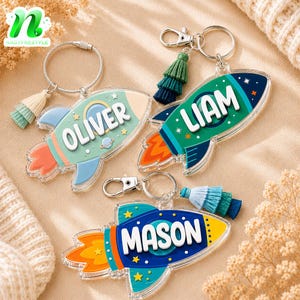 May include: Three acrylic rocket ship keychains featuring the names Oliver, Liam, and Mason in white raised lettering. Each keychain includes a metal ring or clasp, a multi-layered fabric tassel, and colorful space-themed graphics with stars.