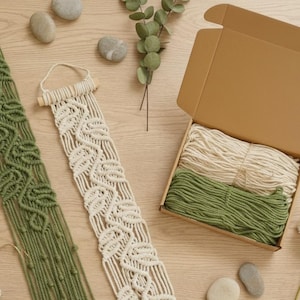 May include: A macrame wall hanging in cream and olive green, with a box of cream and green macrame cord. The wall hangings have a leaf pattern. The box is open, revealing the neatly wound cord. Decorative stones and greenery are also present.