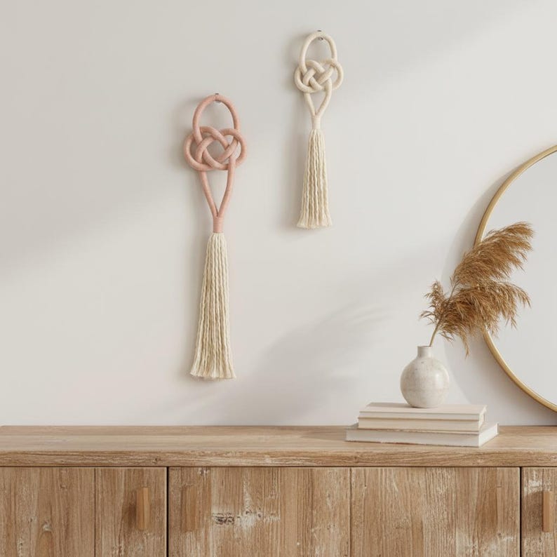May include: Two wall hangings featuring intricate knotted rope designs with long, dangling tassels. One hanging is a soft dusty pink, while the other is a natural cream color. Both are suspended vertically against a plain wall surface.