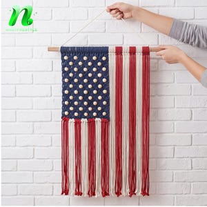 May include: Macrame wall hanging depicting the American flag. The flag features a blue field with wooden bead stars, red and white stripes, and a wooden dowel for hanging. The flag is displayed against a white brick wall.
