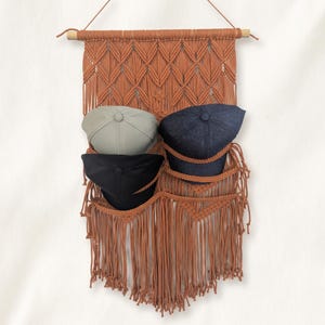 May include: A terracotta-colored macrame wall hanging featuring a wooden dowel at the top and intricate knotted patterns. The piece includes three integrated pockets holding three baseball caps in light gray, dark blue, and black colors.