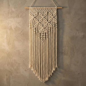 May include: Cream-colored macrame wall hanging. The design features geometric patterns and knotted details, suspended from a wooden dowel. The bottom of the macrame has a triangular shape with long fringe.