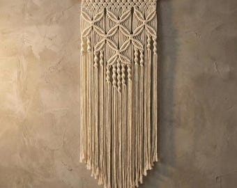 May include: Cream-colored macrame wall hanging. The design features geometric patterns and knotted details, suspended from a wooden dowel. The bottom of the macrame has a triangular shape with long fringe.
