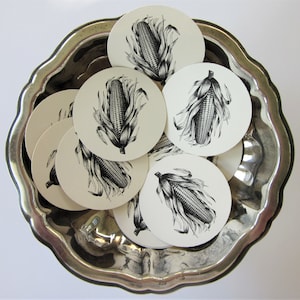 May include: A set of eight white paper coasters with black and white illustrations of corn on the cob. The coasters are arranged in a circular pattern on a silver tray.