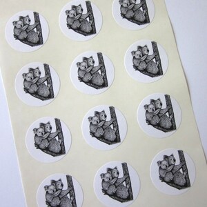 May include: Twelve round stickers featuring a black and white illustration of two koalas hugging on a tree trunk. The stickers are white with a black outline.