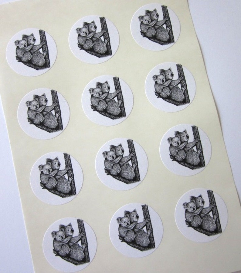 May include: Twelve round stickers featuring a black and white illustration of two koalas hugging on a tree trunk. The stickers are white with a black outline.