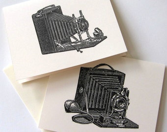 Pode incluir: Duas câmeras vintage com fole e lentes. As câmeras são em preto e branco e têm um estilo de ilustração detalhado. A câmera de cima está aberta e a câmera de baixo está fechada.