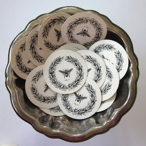 May include: A silver bowl filled with round white paper coasters featuring a black and white illustration of a bee inside a wreath of leaves. The text "Farm to Table" is printed on the coasters.