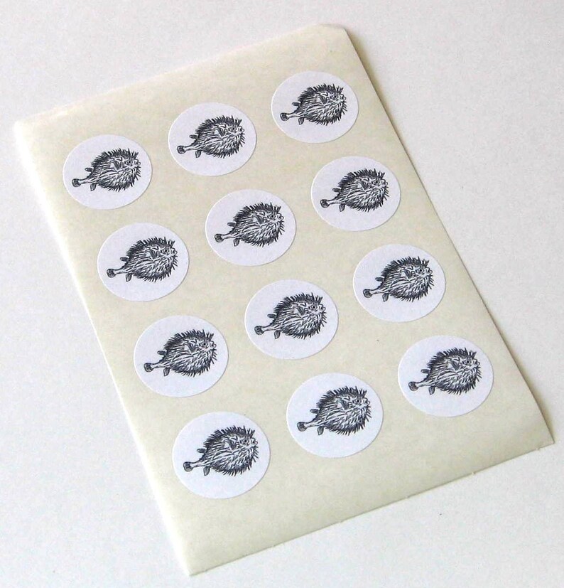May include: A sheet of white sticker paper with 12 black and white illustrations of pufferfish. The stickers are round and have a white background.