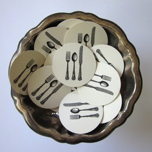 Puede incluir: Una bandeja plateada con borde festoneado contiene una colección de posavasos redondos blancos con ilustraciones de tinta negra de tenedores, cuchillos y cucharas.