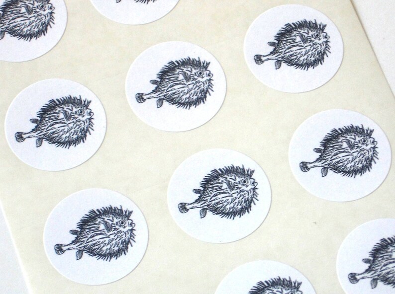 May include: A sheet of round stickers featuring black and white illustrations of pufferfish. Each sticker has a white background and a black outline.