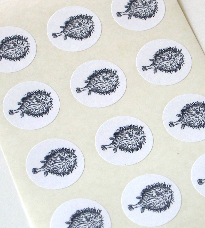 May include: A sheet of white paper with 12 black and white stickers featuring a cartoon pufferfish. Each sticker is round and has a white background.