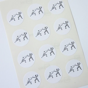 May include: A sheet of white paper with twelve round stickers featuring a black and white illustration of two fencers. Each sticker is approximately 2 inches in diameter.