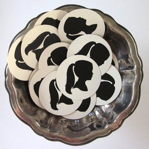 May include: A silver bowl filled with white round paper circles with black silhouette images of people with long hair.