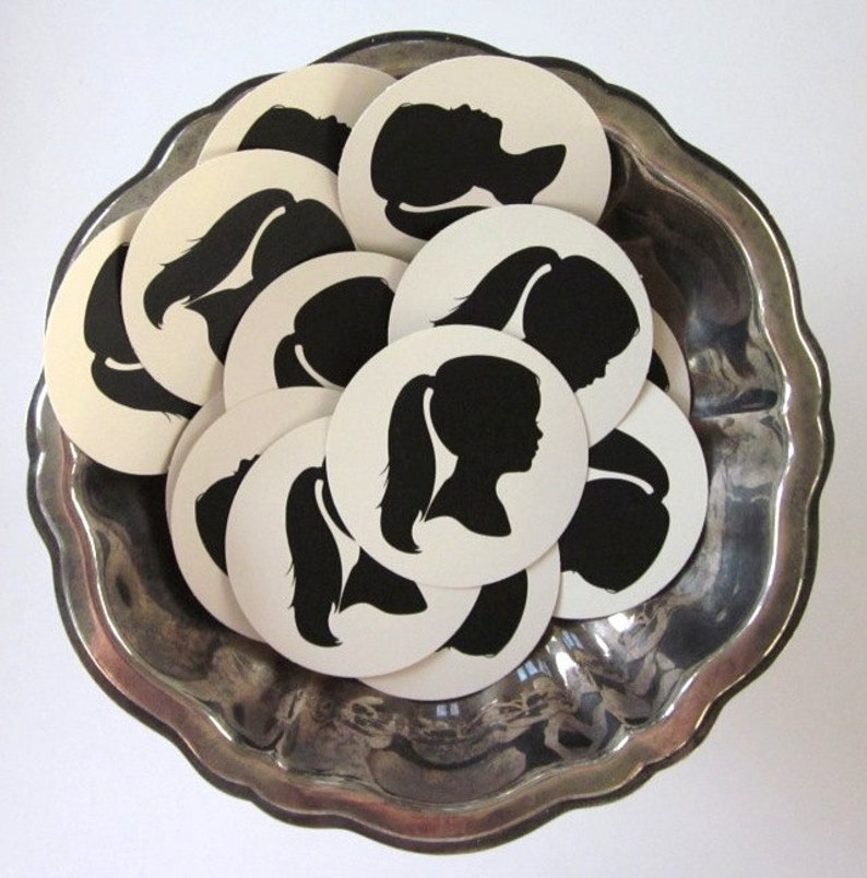 May include: A silver bowl filled with white round paper circles with black silhouette images of people with long hair.
