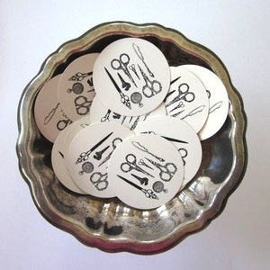 May include: A silver bowl filled with round white paper coasters featuring black and white illustrations of scissors, needles, and thread. The coasters are arranged in a circular pattern.