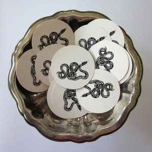 Peut inclure: Un plateau en argent contenant une collection de dessous-de-verre en papier ronds avec des illustrations de serpents en noir et blanc. Les dessous-de-verre sont disposés en cercle.