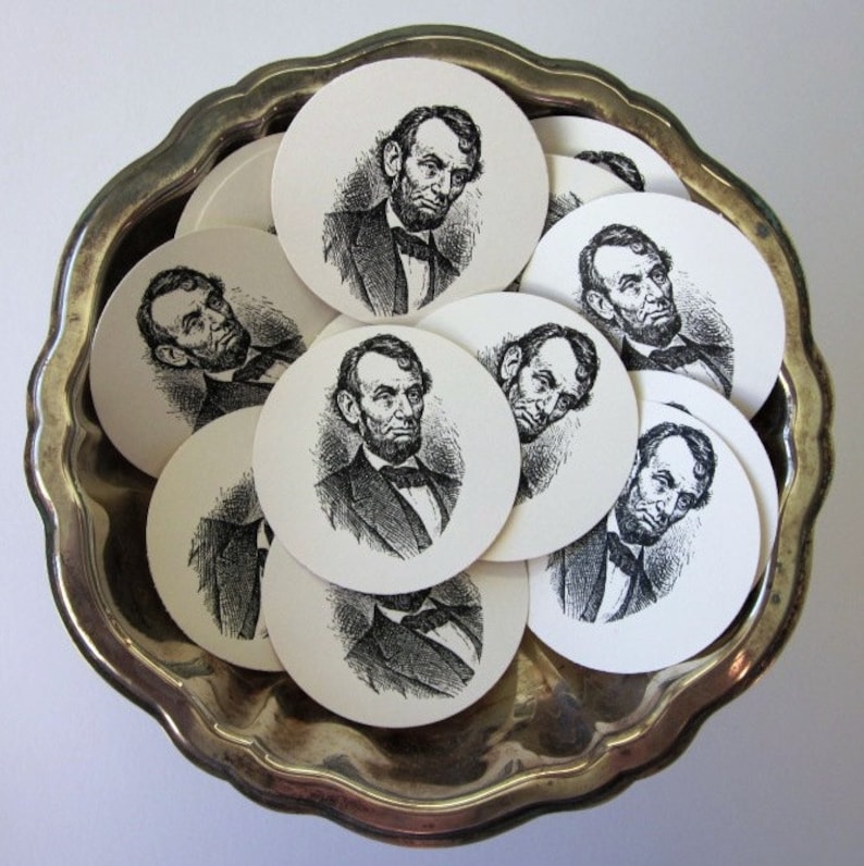 Pu&ograve; includere: Un vassoio d'argento con una collezione di sottobicchieri rotondi di carta con illustrazioni in bianco e nero di Abraham Lincoln. Ogni sottobicchiere presenta un'immagine diversa di Lincoln.