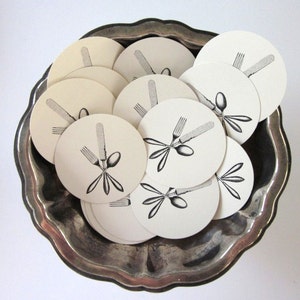 May include: A set of white round paper coasters with black ink illustrations of a fork, knife, and spoon. The coasters are arranged in a silver bowl.