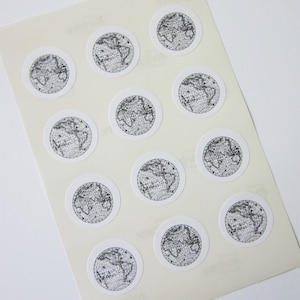 Könnte beinhalten: Ein Blatt weißes Papier mit zwölf schwarzen und weißen Globus-Aufklebern. Jeder Aufkleber zeigt eine Weltkarte im Vintage-Stil.