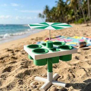 Pode incluir: Uma mesa de praia portátil branca e verde com um poste central, um tampo quadrado com quatro porta-copos circulares e dois compartimentos quadrados, e um guarda-sol a condizer com riscas verdes e brancas no centro.