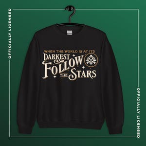 Könnte beinhalten: Schwarzes Sweatshirt mit dem Text "When the world is at its darkest, follow the stars" in einem Vintage-Stil. Das Design umfasst Sterne und ein rundes Emblem. Das Sweatshirt hängt an einem schwarzen Kleiderbügel.