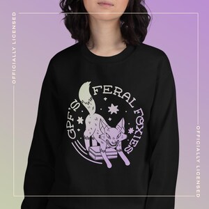 Puede incluir: Sudadera negra con un diseño gráfico circular que presenta un zorro morado y las palabras "GPF'S FERAL FOXIES". El zorro está representado encima de una pila de libros. Las palabras "OFFICIALLY LICENSED" están impresas verticalmente en los lados.