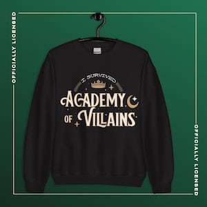 Op de afbeelding: Zwart sweatshirt met de tekst "I Survived Academy of Villains" in een goud en wit ontwerp. Het sweatshirt hangt aan een zwarte hanger tegen een donkergroene achtergrond. De woorden "Officieel gelicentieerd" zijn verticaal gedrukt.