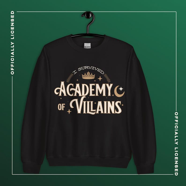 Op de afbeelding: Zwart sweatshirt met de tekst "I Survived Academy of Villains" in een goud en wit ontwerp. Het sweatshirt hangt aan een zwarte hanger tegen een donkergroene achtergrond. De woorden "Officieel gelicentieerd" zijn verticaal gedrukt.