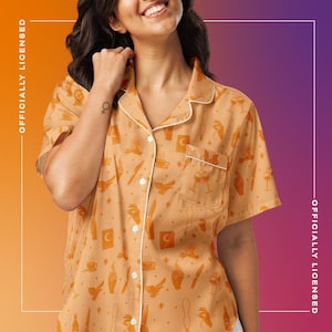 Puede incluir: Camisa de pijama naranja de manga corta con ribete blanco y un bolsillo. La parte superior tiene un estampado de varias ilustraciones naranjas, incluyendo lunas, cuchillos y copas de vino. Las palabras "OFFICIALLY LICENSED" están impresas verticalmente.