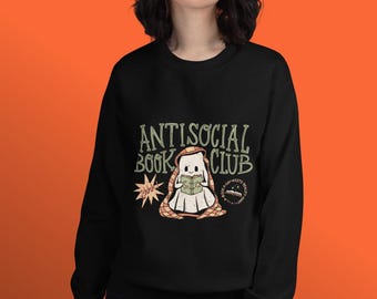 Puede incluir: Sudadera negra con el texto "ANTISOCIAL BOOK CLUB" en verde y blanco. Un fantasma de dibujos animados leyendo un libro está en el centro. Una estrella amarilla y un pequeño gráfico circular también están en la sudadera.