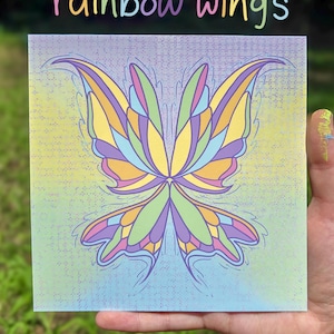 May include: Square canvas art featuring a colorful butterfly wing design. The wings are segmented with shades of yellow, green, purple, and pink. The text "rainbow wings" is written above the artwork in a rainbow gradient.