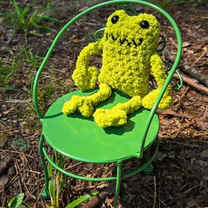 May include: A lime green, plush frog toy with black eyes and a stitched mouth sits on a small, green metal chair. The chair has a round seat and curved back. The frog is made of a fuzzy material. The background is a natural outdoor setting.