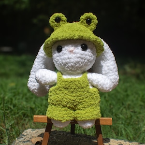 May include: A handmade white crochet bunny wearing a green frog hat and overalls. The bunny has large black eyes and is posed on a small wooden stand against a blurred green background. The hat features two round, dark green eyes.