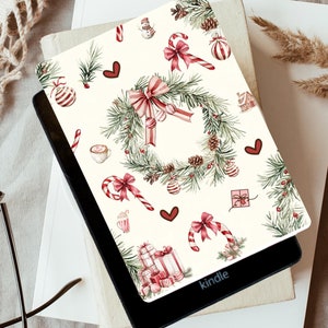 Könnte beinhalten: Ein Kindle E-Reader mit einer festlichen Hülle mit einem Weihnachtskranz, Zuckerstangen und Geschenkboxen auf cremefarbenem Hintergrund. Das Design umfasst rote Schleifen, Ornamente und Tannennadeln, die ein festliches Thema schaffen.