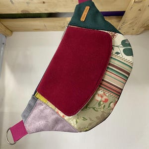 Peut inclure: Sac banane fait main avec un design patchwork. Le sac présente un rabat rouge foncé, un dessus vert et une variété de tissus à motifs dans les tons de rose, vert et beige. Une sangle rose avec un anneau argenté et une étiquette en cuir avec le texte "Mama Zofia" sont visibles.