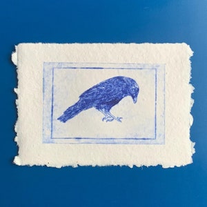 Könnte beinhalten: Ein blauer Druck eines Raben auf strukturiertem weißem Papier mit gezacktem Rand. Der Rabe befindet sich in einem blauen rechteckigen Rahmen. Der Hintergrund ist einfarbig blau.