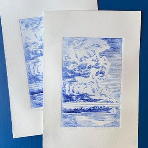 Pode incluir: Duas gravuras de arte paisagística em azul e branco. Cada gravura apresenta uma representação detalhada de uma paisagem de nuvens sobre um corpo de água. As gravuras estão em papel branco, com um fundo azul liso.
